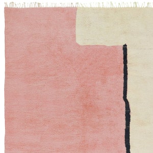 K&ouml;nnte beinhalten: Ein rosafarbener und cremefarbener Teppich mit einem schwarzen geometrischen Muster. Der Teppich hat einen Fransenrand.