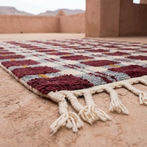 Puede incluir: Una alfombra rectangular con un patr&oacute;n repetitivo de cuadrados burdeos y blancos, acentuados con gris y amarillo. La alfombra tiene una franja de color crema a lo largo de un borde. La alfombra est&aacute; hecha de un material grueso y tejido.