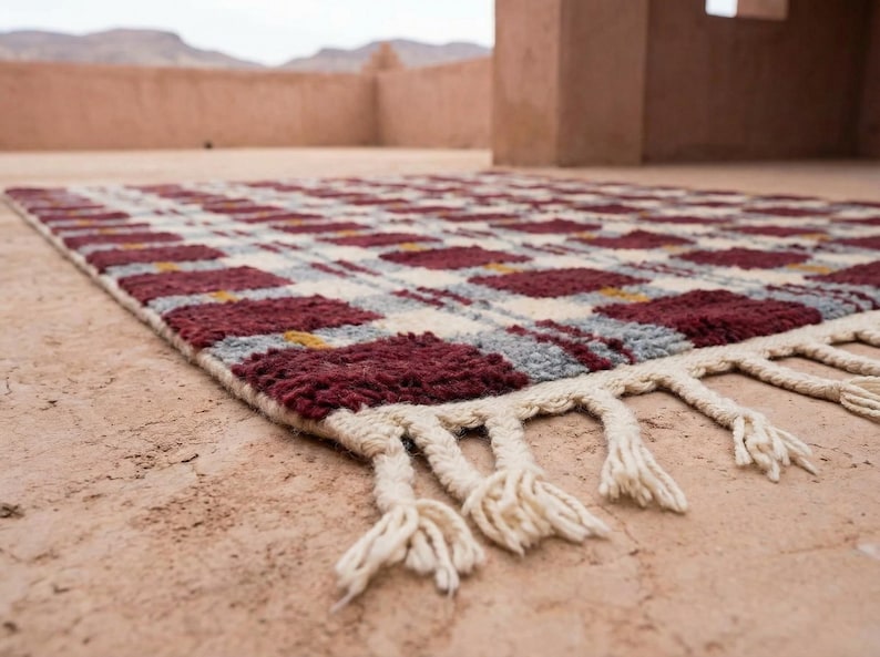Puede incluir: Una alfombra rectangular con un patr&oacute;n repetitivo de cuadrados burdeos y blancos, acentuados con gris y amarillo. La alfombra tiene una franja de color crema a lo largo de un borde. La alfombra est&aacute; hecha de un material grueso y tejido.