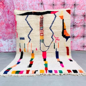May include: A white wool rug with a geometric pattern in shades of blue, red, yellow, green, and pink. The rug has a fringe border.
