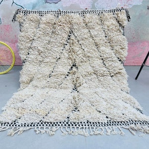 May include: A white shag rug with a black and white geometric pattern border. The rug has a fringe border and is made of soft, fluffy fibers.