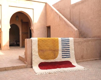 Puede incluir: Alfombra rectangular blanquecina con diseño geométrico. Presenta una gran forma amarilla, una forma roja y rayas azules. La alfombra se exhibe al aire libre contra un edificio color melocotón. Medidas en centímetros.