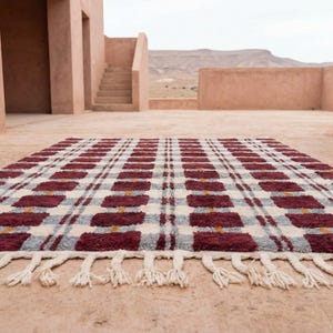 Puede incluir: Una alfombra rectangular con un patr&oacute;n de cuadros en burdeos, gris y crema. La alfombra tiene flecos en el borde inferior. El fondo muestra un edificio con escaleras y una cordillera.