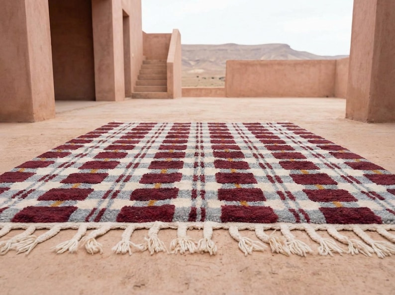 Puede incluir: Una alfombra rectangular con un patr&oacute;n de cuadros en burdeos, gris y crema. La alfombra tiene flecos en el borde inferior. El fondo muestra un edificio con escaleras y una cordillera.