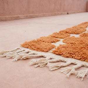 May include: A close-up of a handmade rug with a geometric pattern. The rug features a textured, orange pile with a cream-colored border and fringe. The rug is placed on a pink surface.