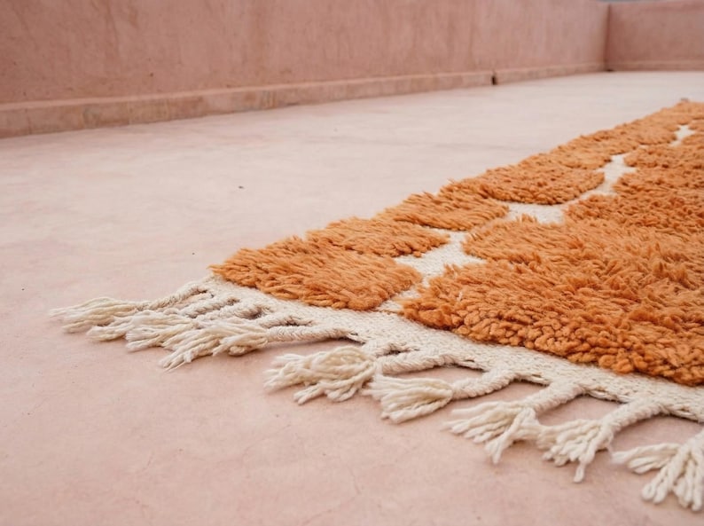 May include: A close-up of a handmade rug with a geometric pattern. The rug features a textured, orange pile with a cream-colored border and fringe. The rug is placed on a pink surface.