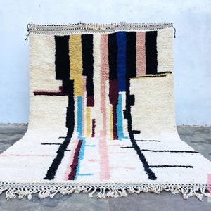 May include: A white, handwoven rug with a geometric pattern in black, blue, pink, yellow, and red. The rug has a fringe border.