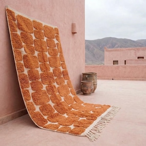 May include: A large, rectangular rug with a repeating pattern of orange squares on a cream background. The rug has a fringed edge and is leaning against a pink wall. The scene is outdoors, with mountains in the background.