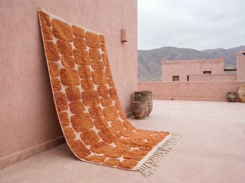 May include: A large, rectangular rug with a repeating pattern of orange squares on a cream background. The rug has a fringed edge and is leaning against a pink wall. The scene is outdoors, with mountains in the background.