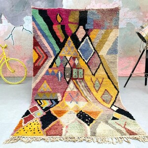 May include: A colorful abstract geometric patterned rug with a fringe border. The rug features a mix of yellow, pink, blue, green, and black colors.