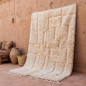 May include: A large, off-white rug with a geometric pattern leans against a textured, reddish-brown wall. The rug has a thick, shaggy texture and a fringe along the bottom edge. The rug's design features a series of rectangular shapes.
