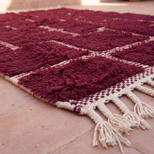 May include: A burgundy area rug with a geometric pattern of squares and lines in off-white. The rug has a thick, textured pile and a fringe border. The rug is made of a soft material and is suitable for a living room or bedroom.