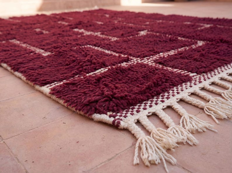 May include: A burgundy area rug with a geometric pattern of squares and lines in off-white. The rug has a thick, textured pile and a fringe border. The rug is made of a soft material and is suitable for a living room or bedroom.