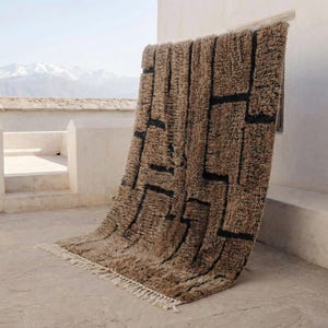 May include: A brown and black geometric patterned rug with a shaggy texture. The rug is displayed outdoors, leaning against a white wall. The design features black rectangular shapes on a brown background. The rug has a fringe along the bottom edge.