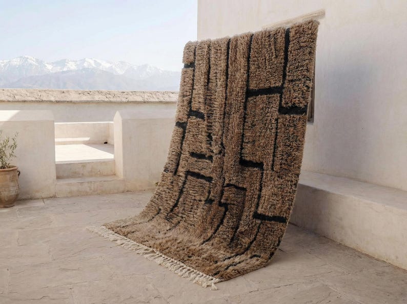 May include: A brown and black geometric patterned rug with a shaggy texture. The rug is displayed outdoors, leaning against a white wall. The design features black rectangular shapes on a brown background. The rug has a fringe along the bottom edge.