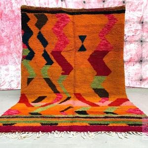 May include: A colorful Moroccan rug with a geometric pattern in shades of orange, red, green, and black. The rug has a fringe border and is made of wool.