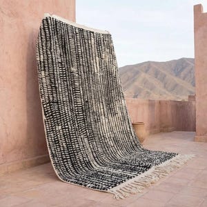 May include: A black and white patterned rug with a fringe. The rug is leaning against a pink wall on a tiled patio. The pattern consists of vertical lines. Mountains are visible in the background.