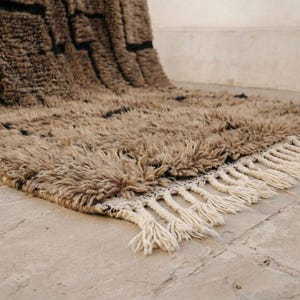 May include: A beige and black Moroccan-style rug with a shaggy texture. The rug features a geometric pattern in black and a fringe border. The rug is made of a soft, plush material.