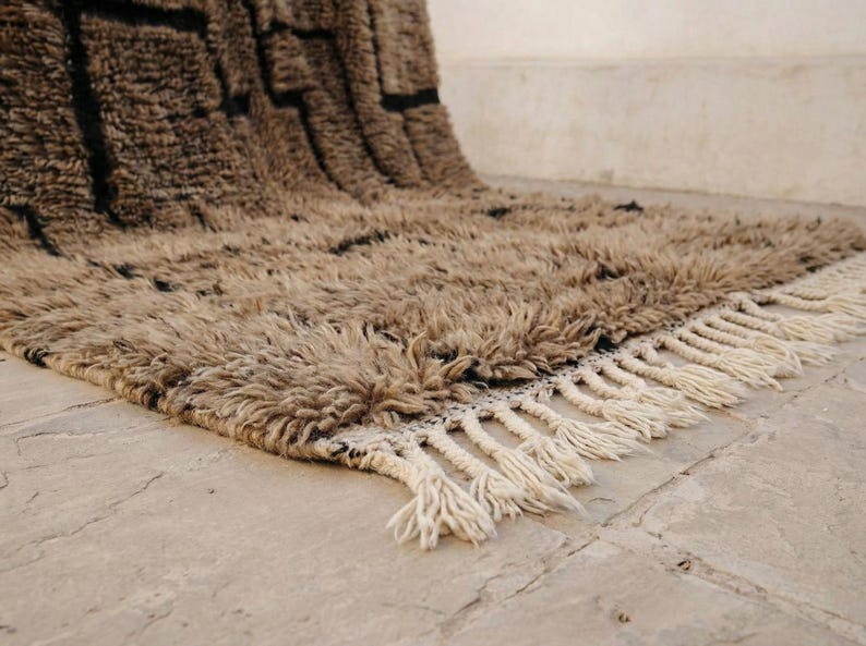 May include: A beige and black Moroccan-style rug with a shaggy texture. The rug features a geometric pattern in black and a fringe border. The rug is made of a soft, plush material.