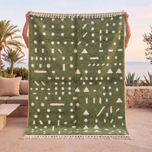 May include: A rectangular area rug featuring a deep olive green background with a repeating pattern of white geometric shapes, including circles, triangles, and dashes. The rug has a thick, shaggy pile texture and white fringe along the top and bottom edges.