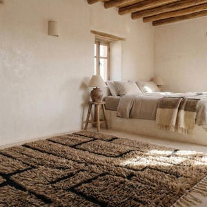May include: A bedroom with a neutral color palette. A large, shaggy brown rug with black geometric patterns sits on the floor. A bed with beige bedding and a wooden side table with a lamp are visible.