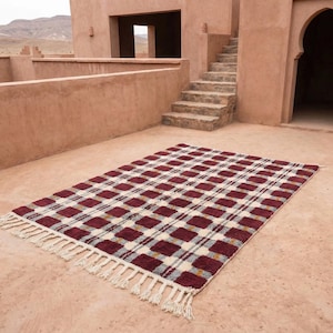 Puede incluir: Una alfombra rectangular con un patr&oacute;n de cuadros en tonos burdeos, crema y gris. La alfombra tiene flecos en los extremos cortos. La alfombra est&aacute; colocada sobre una superficie de color terracota, con un edificio y escaleras al fondo.