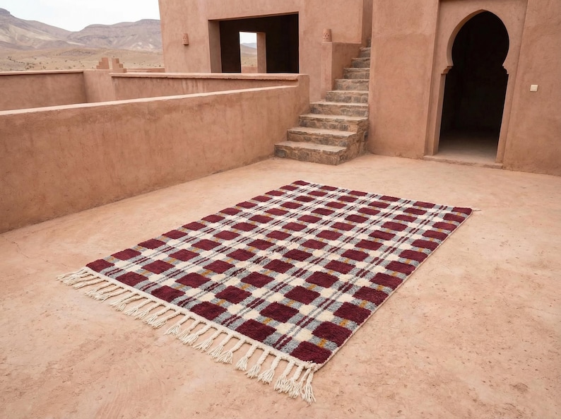 Puede incluir: Una alfombra rectangular con un patr&oacute;n de cuadros en tonos burdeos, crema y gris. La alfombra tiene flecos en los extremos cortos. La alfombra est&aacute; colocada sobre una superficie de color terracota, con un edificio y escaleras al fondo.