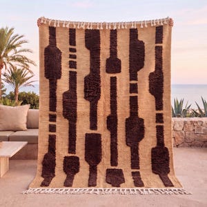 May include: A rectangular area rug featuring a tan base with a repeating pattern of dark brown, shaggy, vertical geometric shapes. The rug has a textured weave and is finished with short, cream-colored fringe tassels along the top and bottom edges.