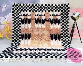May include: A large, rectangular Berber rug with a geometric design. The rug features a black and white checkered border, with a central pattern of black, white, and tan diamond shapes. The rug has a fringed edge and is displayed on a light-colored surface.