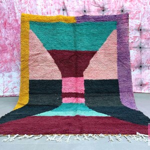 May include: A colorful geometric rug with a pink, teal, burgundy, black, and purple design. The rug has a fringe border and is made of wool.