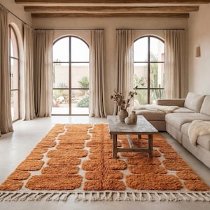 May include: An orange and white patterned rug with a fringed edge is the focal point of a living room. The rug is placed on a light-colored floor, with a beige sectional sofa and a wooden coffee table. Large arched windows with beige curtains are in the background.