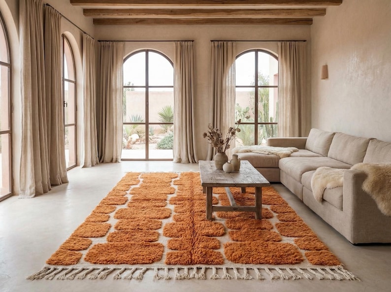 May include: An orange and white patterned rug with a fringed edge is the focal point of a living room. The rug is placed on a light-colored floor, with a beige sectional sofa and a wooden coffee table. Large arched windows with beige curtains are in the background.