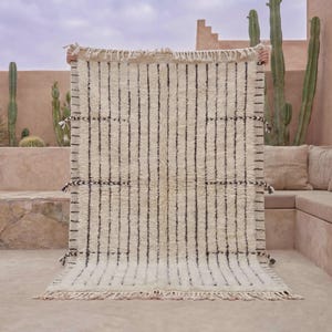 May include: A rectangular off-white area rug featuring a series of vertical black stripes. The rug has a thick, textured pile and is finished with knotted fringe along the top and bottom edges, with small decorative tassels along the sides.