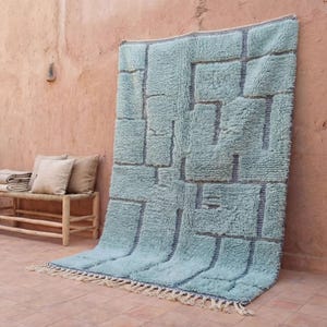 May include: A large, rectangular, light blue rug with a geometric pattern of darker blue rectangles. The rug has a shaggy texture and a fringe along the bottom edge. It is leaning against a textured, reddish-brown wall.