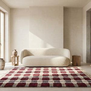 Puede incluir: Un sal&oacute;n con un sof&aacute; curvo color crema y una alfombra a cuadros burdeos y crema. La habitaci&oacute;n tiene paredes neutras, una mesa auxiliar de madera y una ventana grande. Tambi&eacute;n hay jarrones decorativos y un taburete de madera.