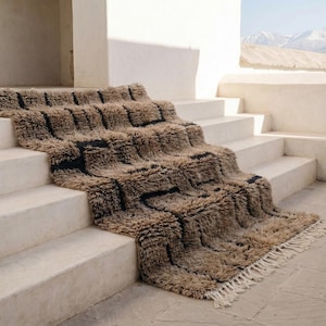 May include: A textured, rectangular rug with a geometric pattern of brown and black squares cascading down a set of white steps. The rug has a shaggy texture and a fringe along the bottom edge. The scene is outdoors with a mountain range in the background.