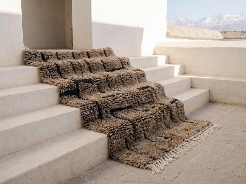May include: A textured, rectangular rug with a geometric pattern of brown and black squares cascading down a set of white steps. The rug has a shaggy texture and a fringe along the bottom edge. The scene is outdoors with a mountain range in the background.