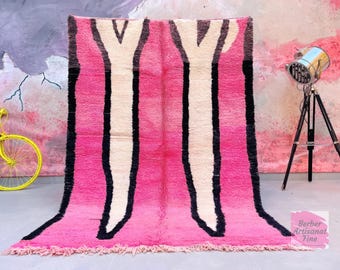 May include: A vibrant pink rug with a bold design. The rug features a central pattern of white and black vertical shapes against a pink background. The rug's edges are fringed, adding texture. A decorative lamp and a yellow bicycle are in the background.