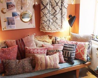Puede incluir: Un sofá cama de madera con una variedad de almohadas de colores con patrones. Las almohadas están dispuestas en un estilo casual y bohemio. El sofá cama está contra una pared con una alfombra de color crema colgada.