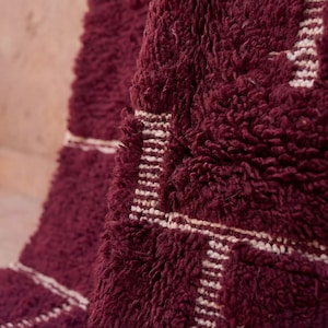 May include: Close-up of a burgundy rug with a thick, textured pile. The rug features a geometric design in off-white, with rectangular and linear patterns. The rug's plush texture and rich color create a warm, inviting aesthetic.