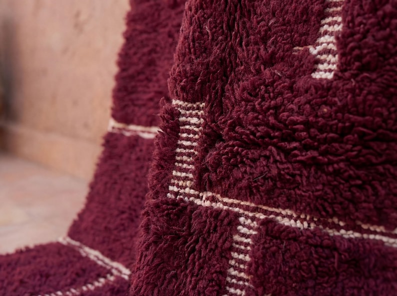 May include: Close-up of a burgundy rug with a thick, textured pile. The rug features a geometric design in off-white, with rectangular and linear patterns. The rug's plush texture and rich color create a warm, inviting aesthetic.