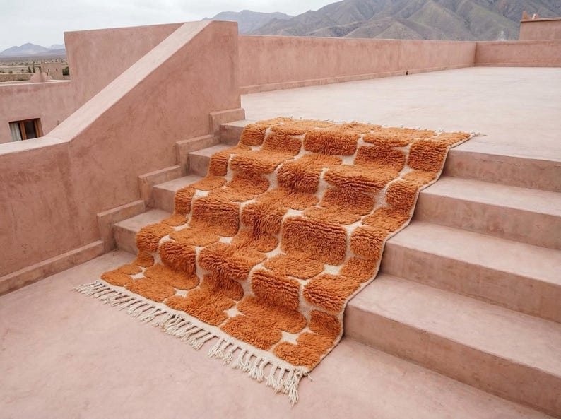 May include: An orange and cream-colored rug with a geometric pattern is draped over a set of stairs. The rug has a shaggy texture and a fringe along the bottom edge. The stairs and surrounding architecture are a muted peach color.