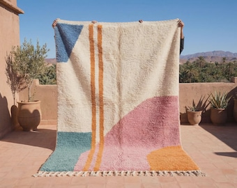Puede incluir: Una alfombra rectangular grande de color crema con textura de pelo largo. La alfombra presenta formas geométricas en azul, verde azulado, rosa y naranja, incluyendo dos rayas verticales naranjas en el centro y flecos en el borde inferior.