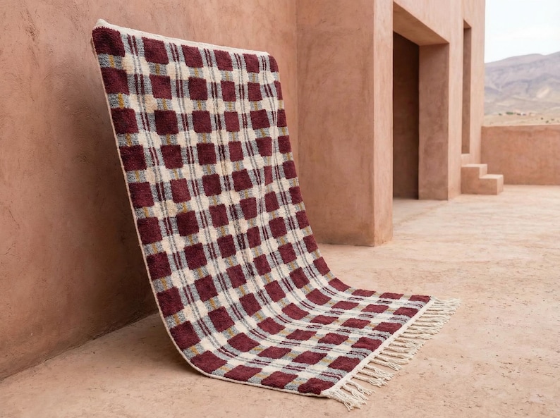 Puede incluir: Una alfombra rectangular tejida con un patr&oacute;n repetido de cuadrados burdeos sobre un fondo crema. La alfombra tiene un borde con flecos y est&aacute; apoyada contra una pared. La alfombra mide aproximadamente 152 cm de largo y 102 cm de ancho.