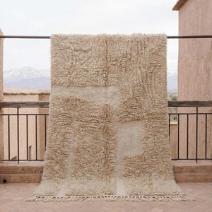 May include: A beige, rectangular rug with a textured, shaggy pile. The rug features a patchwork design with lighter beige sections. It is hanging on a metal rod on a balcony, with a mountain range visible in the background.