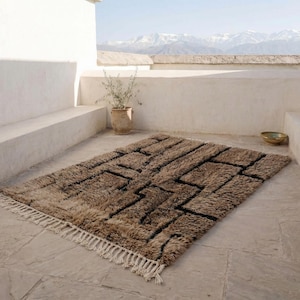 May include: A rectangular brown rug with a geometric black design and white fringe. The rug is placed on a stone patio with a view of mountains in the background. A potted plant and a bowl are also visible.
