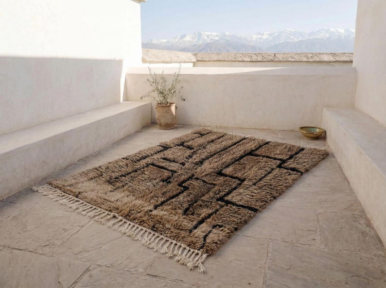 May include: A rectangular brown rug with a geometric black design and white fringe. The rug is placed on a stone patio with a view of mountains in the background. A potted plant and a bowl are also visible.