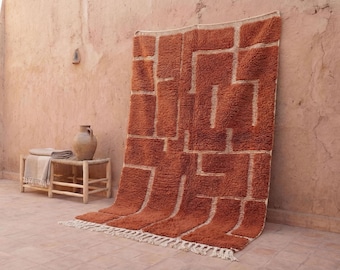 May include: A large, rust-colored rug with a geometric pattern of white lines. The rug is displayed against a textured, peach-colored wall. A small wooden table with a vase and blanket is in the background.