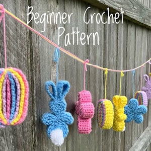 May include: A string of crocheted Easter decorations hanging from a wooden fence. The decorations include a striped egg, a bunny, a cross, a flower, and a yellow chick. The string is made of pink and yellow yarn. The text "Beginner Crochet Pattern" is written above the decorations.