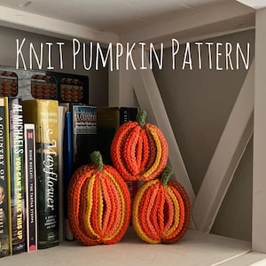 Może przedstawiać: Trzy ręcznie robione na drutach dynie w odcieniach pomarańczu, żółci i brązu z zielonymi ogonkami, ustawione na półce. Tekst KNIT PUMPKIN PATTERN jest widoczny u góry. Dynie mają prążkowaną fakturę i są umieszczone przed książkami.